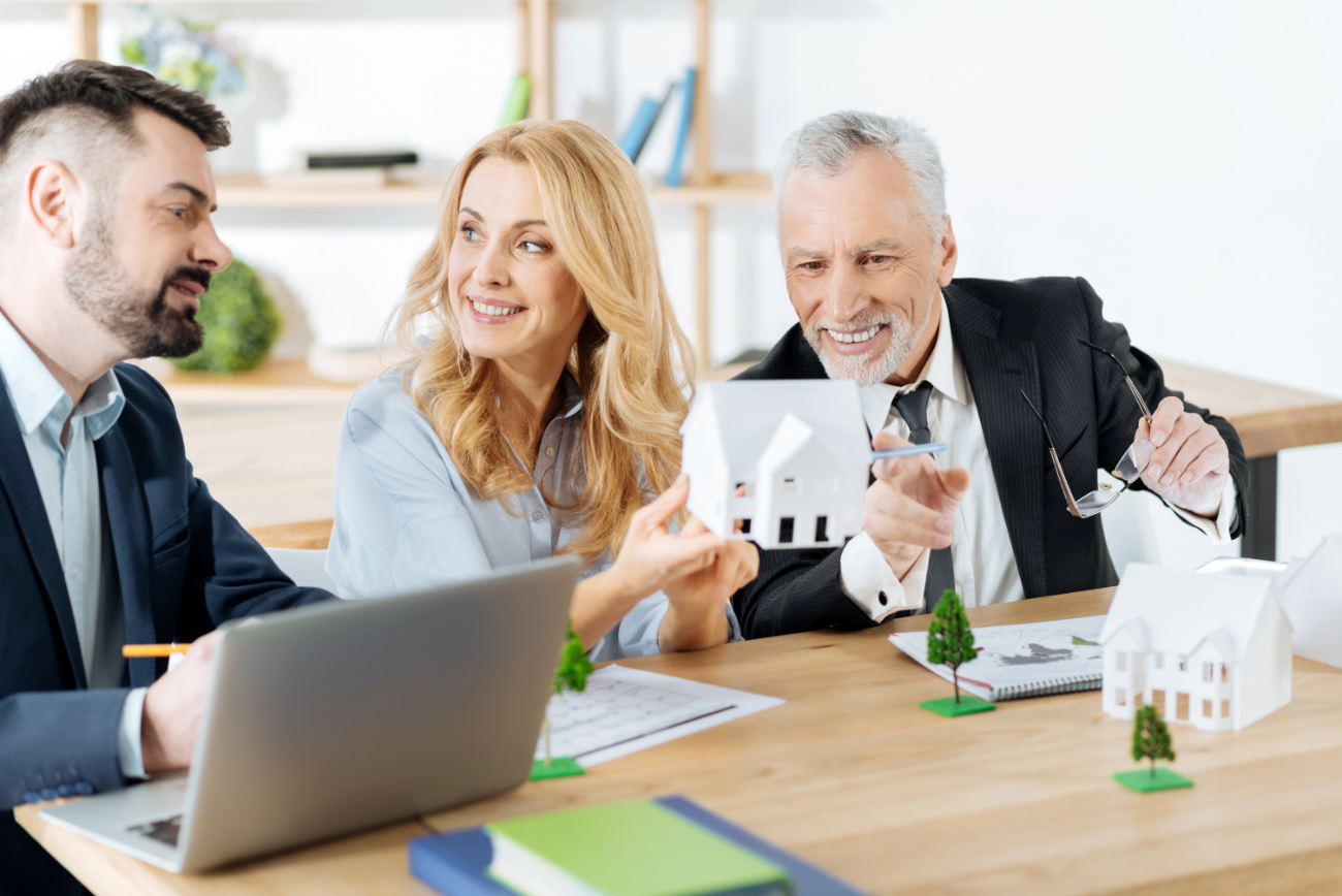 Cheerful realtor looking at her colleague while holding a miniature house